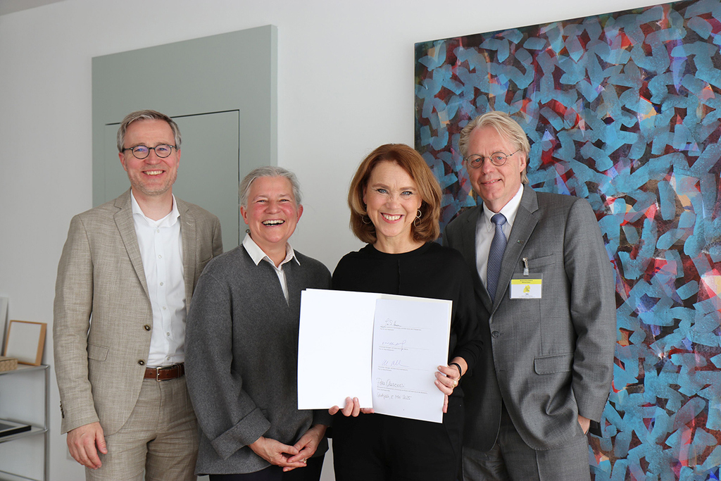 Vier Personen, eine hält ein unterzeichnetes Dokument in der Hand, vor einem bunten Kunstwerk.